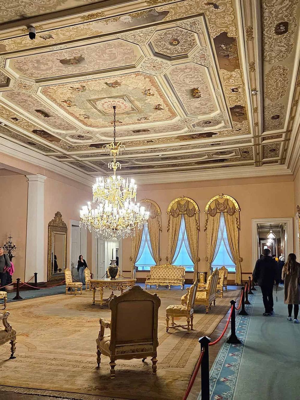 Elegant mansion interior with ornate ceiling chandeliers and luxurious gold and cream furnishings.