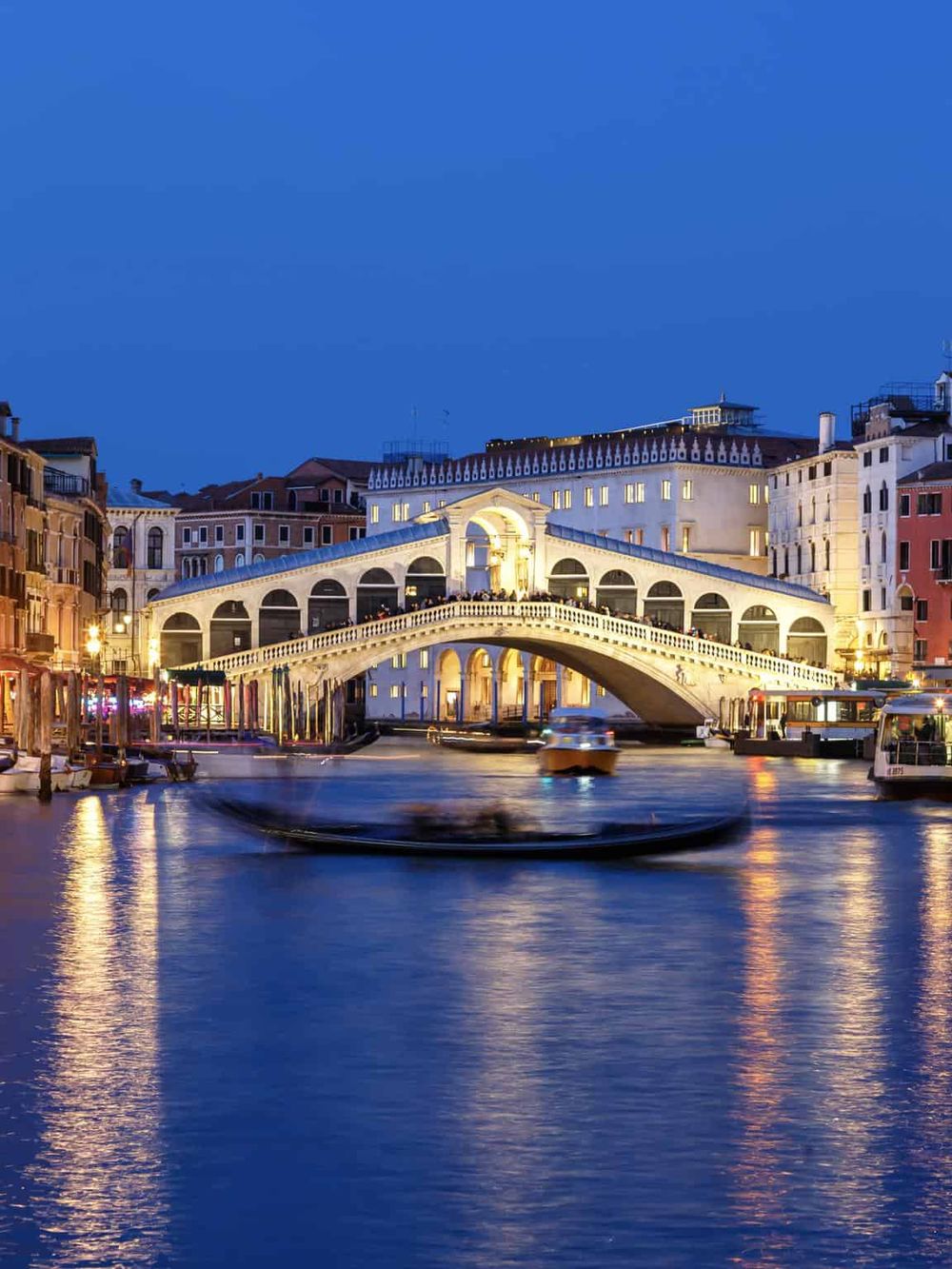 Historic Venice Rialto Bridge illuminated at night with water taxis and reflections.
