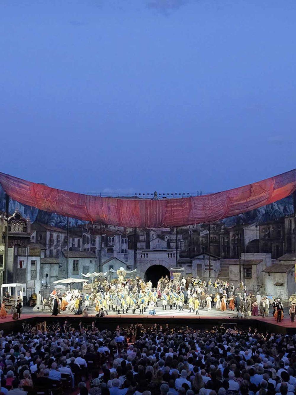 Colorful outdoor theatrical performance in front of a large audience, showcasing a vibrant stage with elaborate set design.