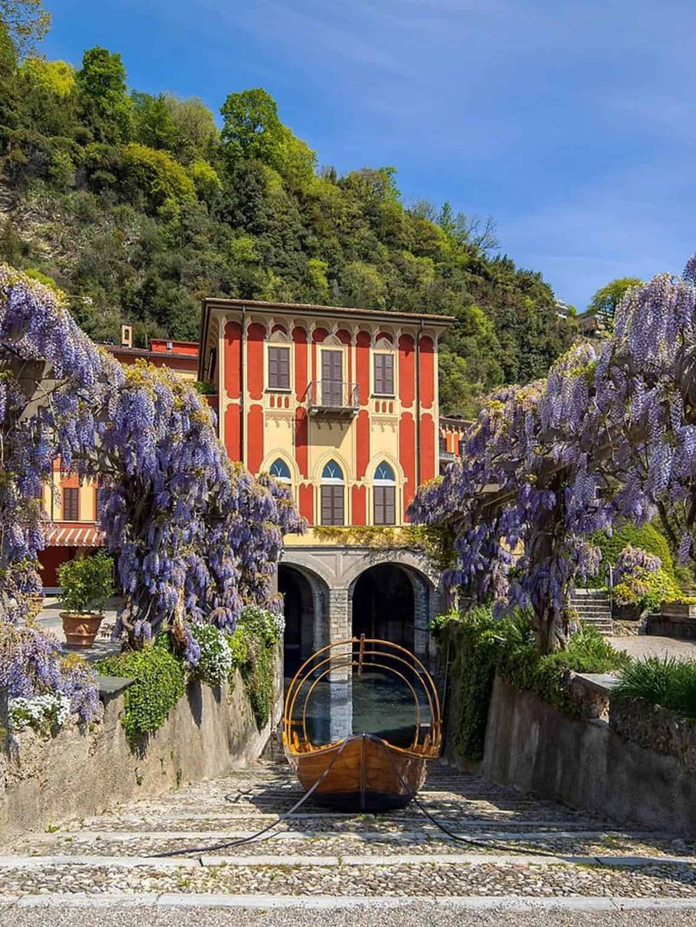 Colorful lakeside villa with blooming wisteria and boat dock, scenic Italian architecture, and lush greenery.