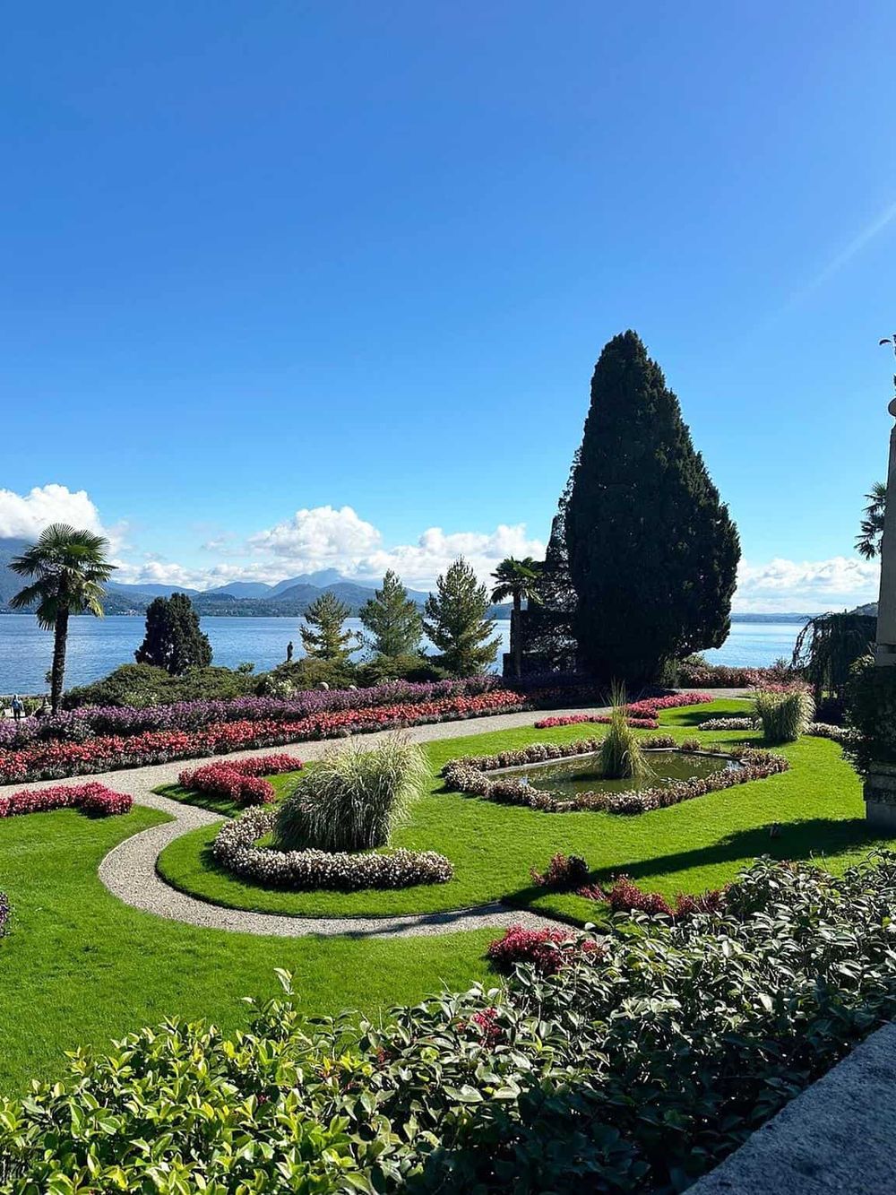 Serene lakeside garden with vibrant flowers, lush greenery, and scenic mountain views under a bright blue sky.