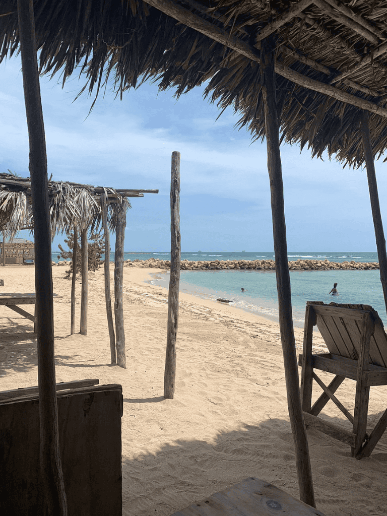 Beach hut with wooden furniture, sandy shore, and calm ocean waves on a bright day.