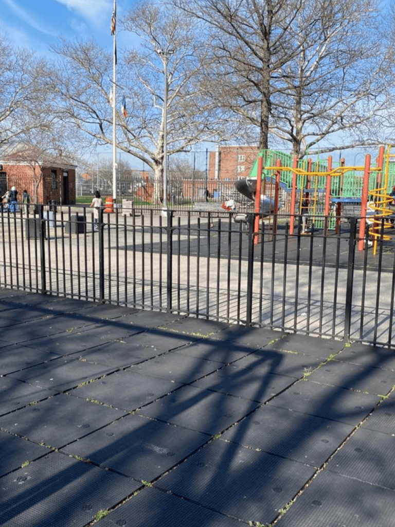 Playground with a black metal fence, slide, kids, and trees on a sunny day.