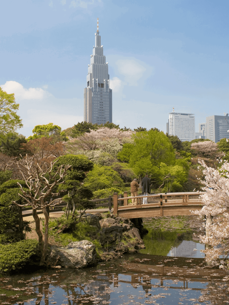 City skyline with skyscraper and park scene with people walking on bridge, spring blossoms, and lush greenery.