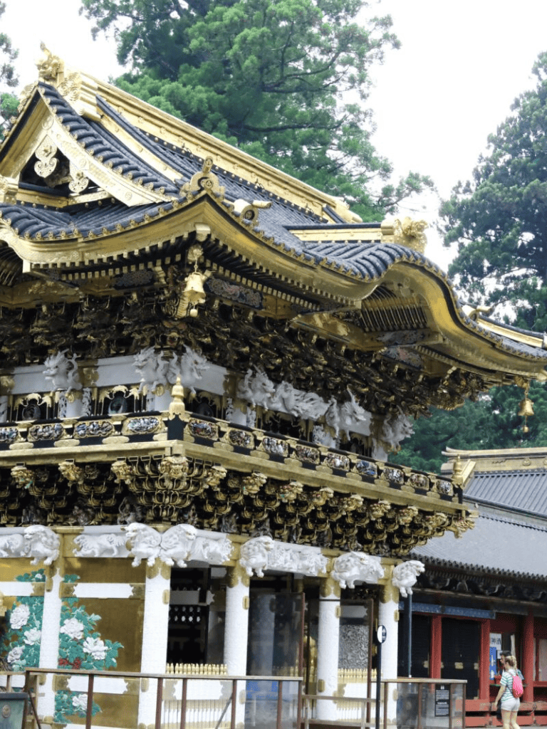 Intricate Japanese temple architecture with gold and black accents, surrounded by lush green trees.