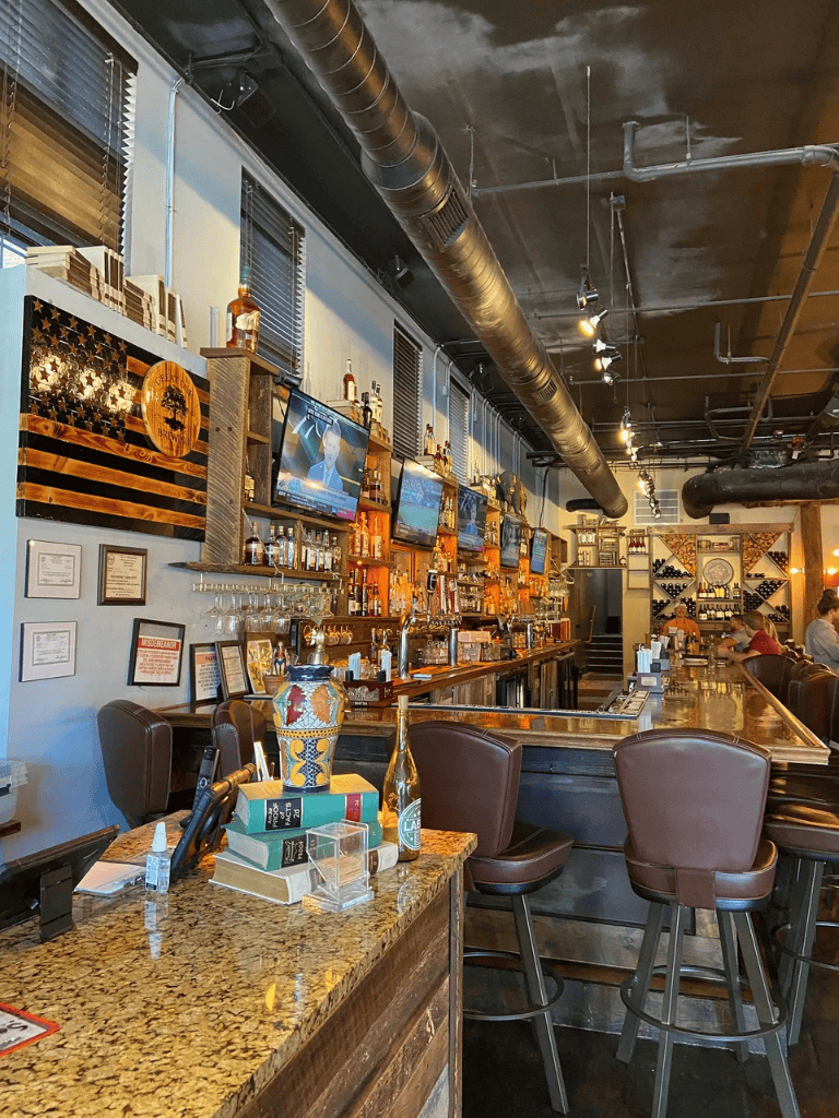 Modern bar with wine racks and multiple TV screens, warm lighting, and a cozy atmosphere.