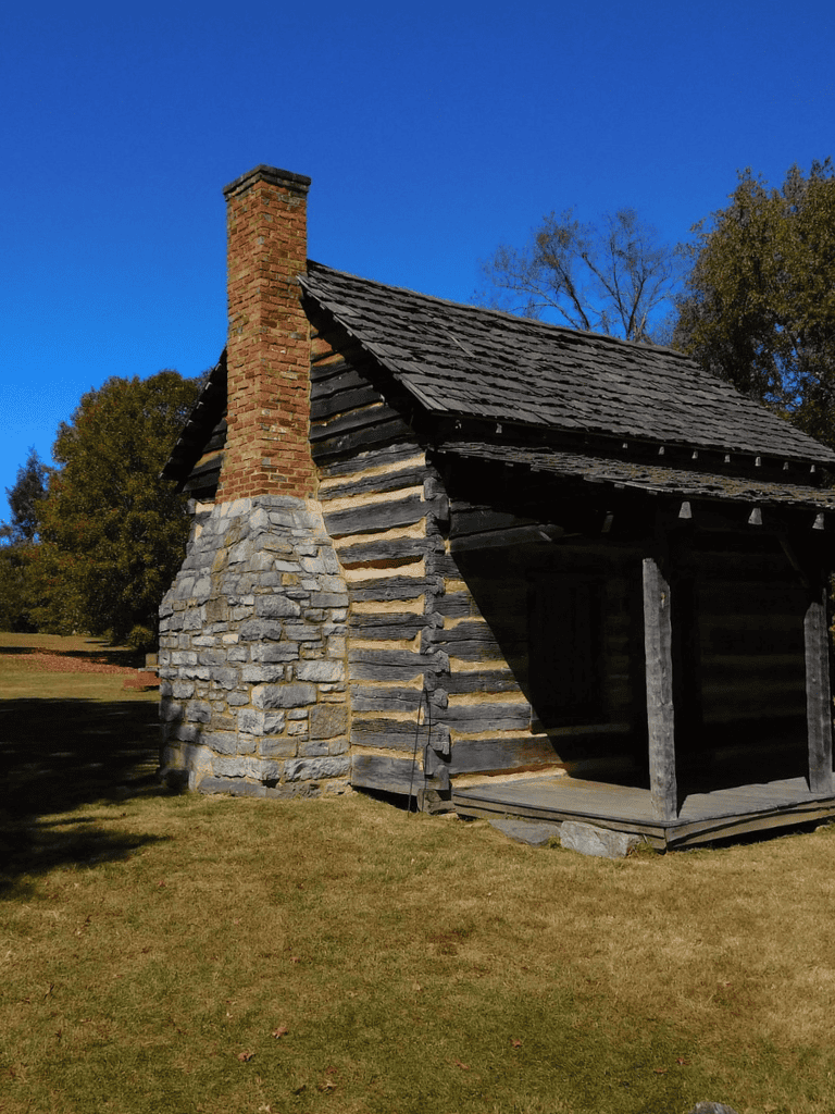 Rustic log cabin with stone chimney on green lawn, historic countryside setting.