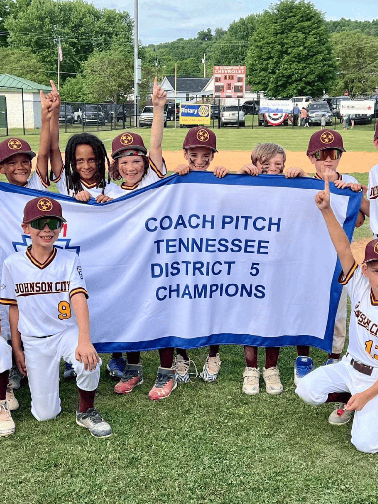 Youth baseball team celebrating championship victory with banner, smiling kids in uniforms, sunny sports field, Tennessee district 5 champs.