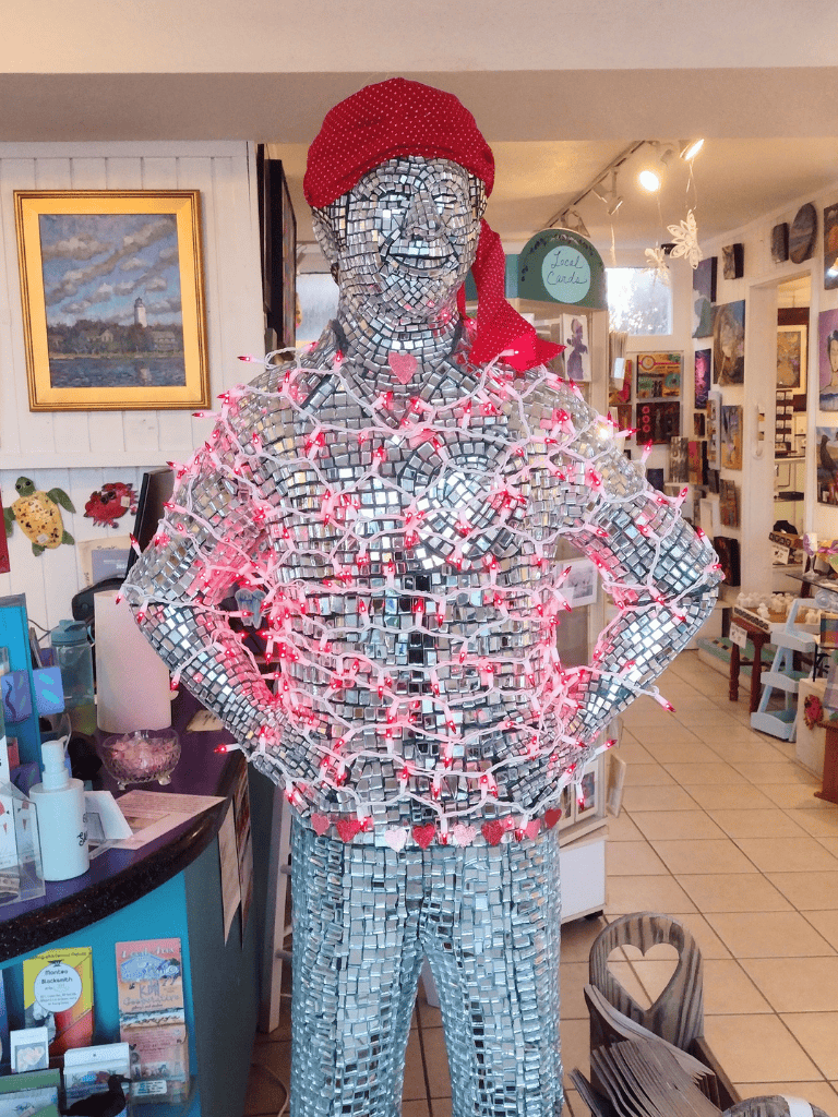 Shiny mirror mosaic statue of man in a store, decorated with red heart lights and a red bandana, inside a gift shop.