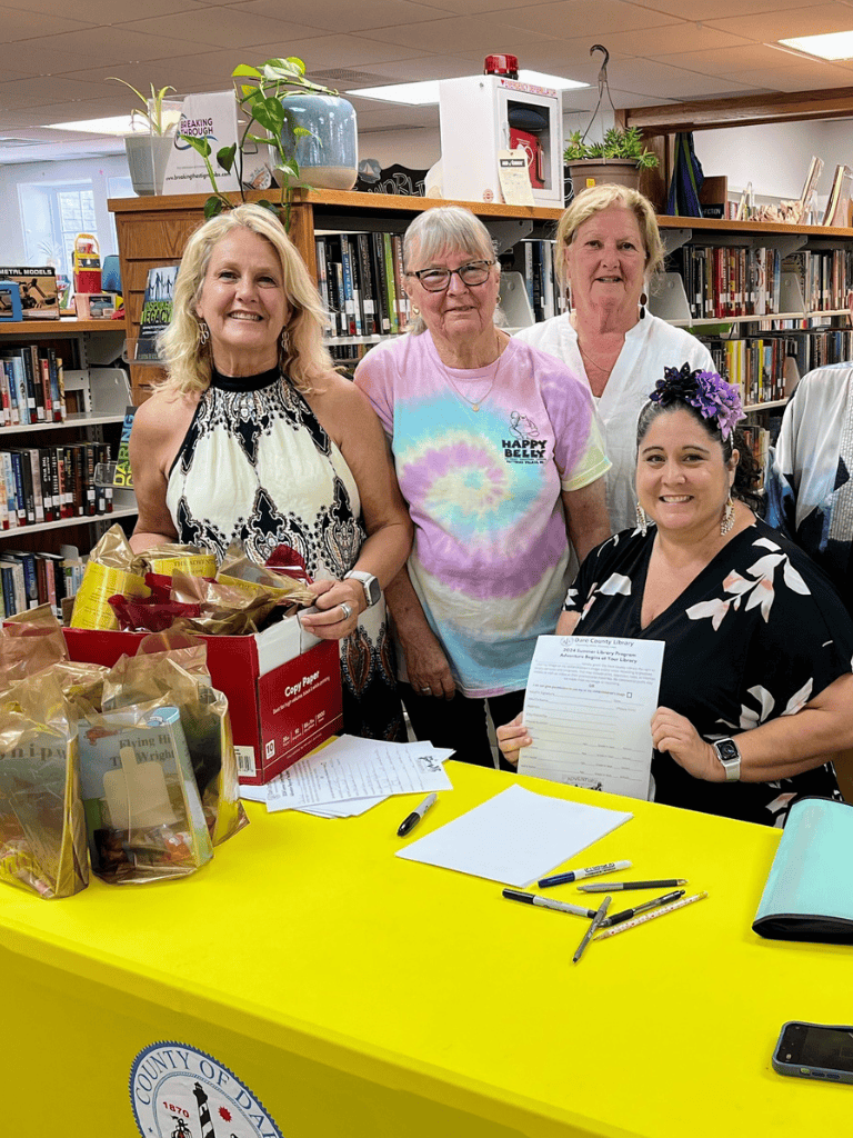 Brightly lit library event with smiling women handing out library materials and signing documents, promoting community engagement and library services.