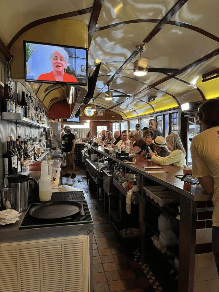 Aerial view of a bustling bar with patrons, bartenders, and a TV screen showing a woman speaking, inside a cozy restaurant.