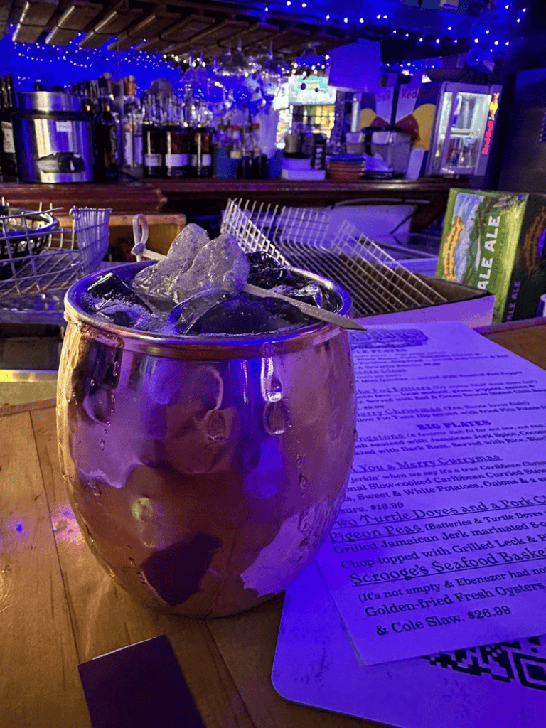 Refreshing cocktail with ice in a metallic glass, bar setting with illuminated decor in the background.