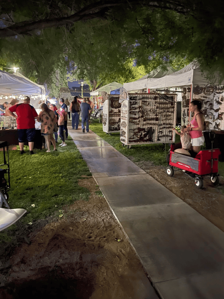 Colorful outdoor night market with tents, shoppers, and street vendors under green tree canopy.
