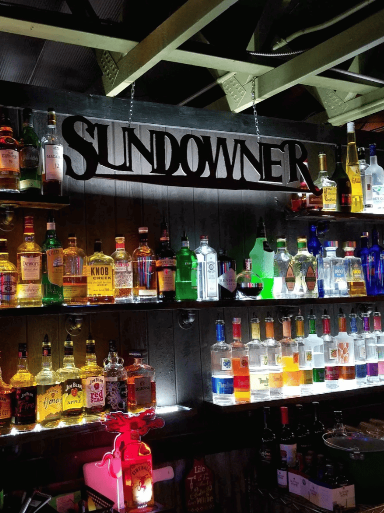 Brightly lit liquor bottles display at Sundowner bar, offering a wide selection for nightlife and cocktail enthusiasts.