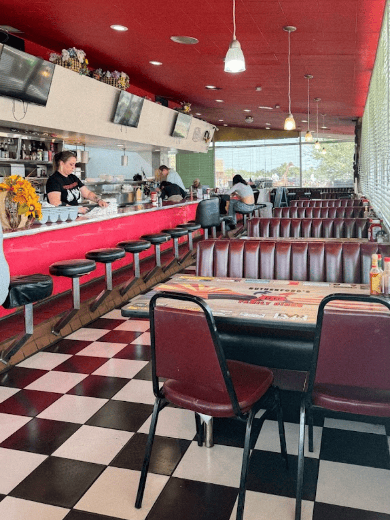 Bright diner interior with checkered floor, red accents, and patrons dining while food is prepared behind the counter.