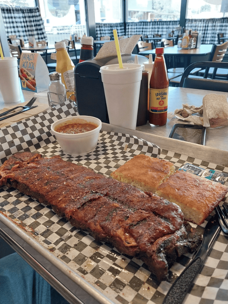BBQ ribs with cornbread and baked potato, served with hot sauce in a casual restaurant setting.
