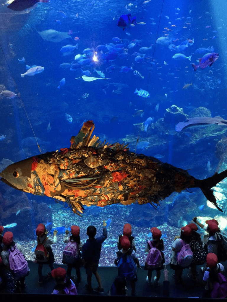 Vibrant underwater scene with diverse fish and a large artistic fish sculpture at an aquarium.