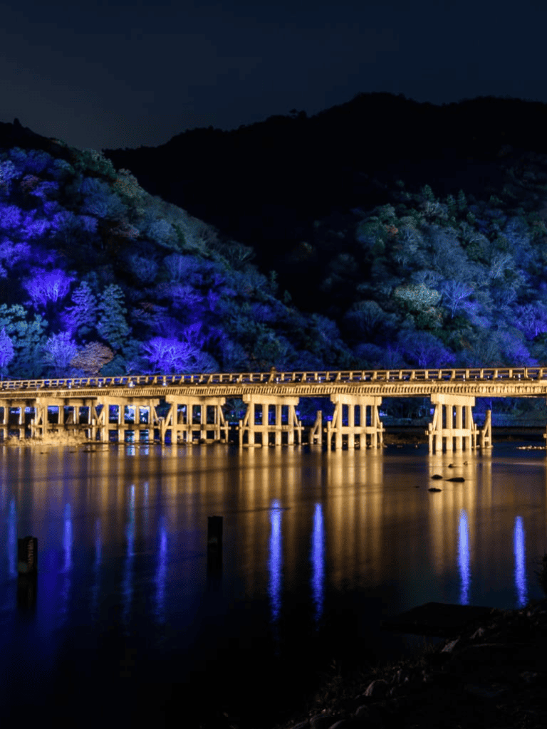 Illuminated bridge over water at night across colorful forested hillside, scenic sunset view.