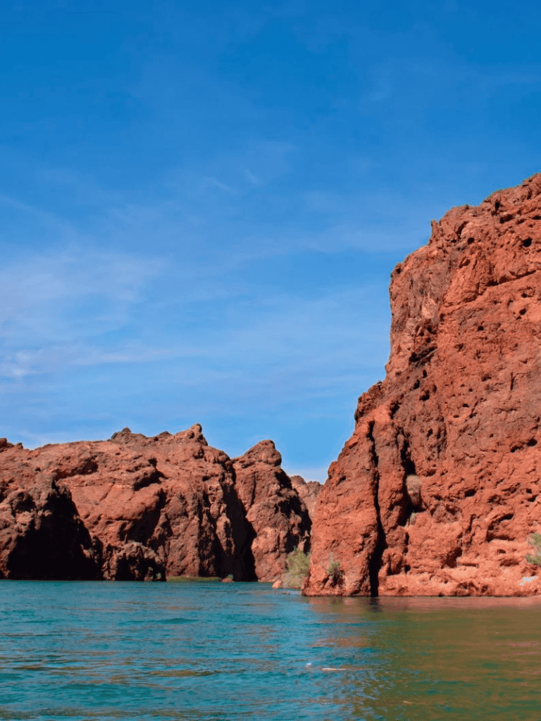 Vivid red canyon cliffs beside calm turquoise water under a bright blue sky.