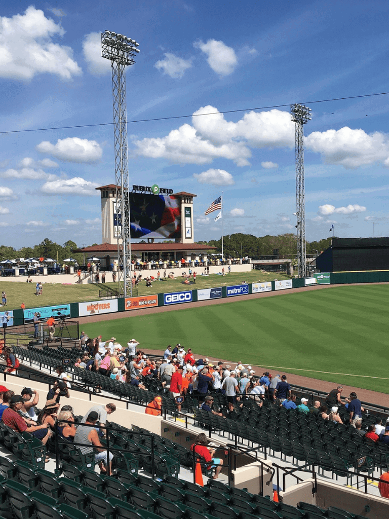Industrial baseball stadium with fans, scoreboard, and American flags on a sunny day.