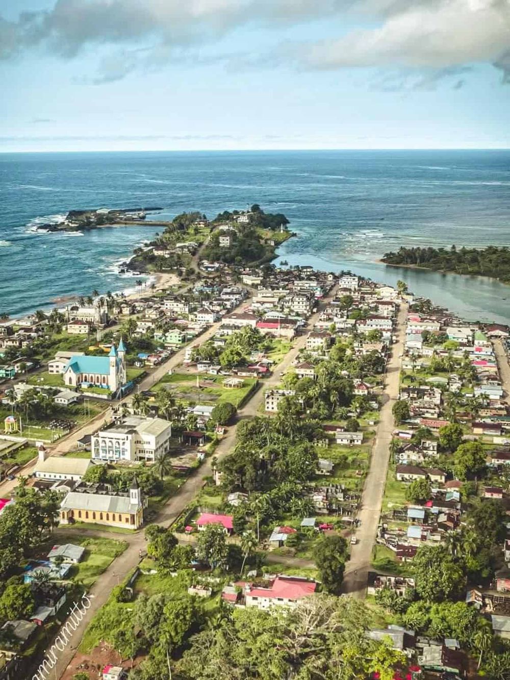 Aerial view of a coastal town with colorful buildings, lush greenery, and the sea in the background.