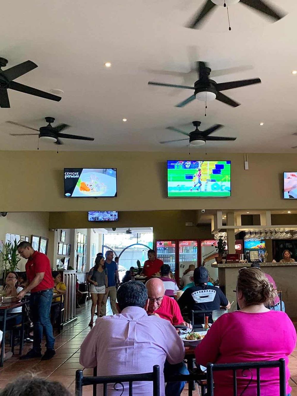 Bright restaurant interior with ceiling fans, multiple TV screens, and diverse diners enjoying meals.