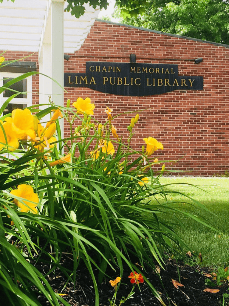 Lima Public Library sign on brick building surrounded by yellow flowers and green foliage.