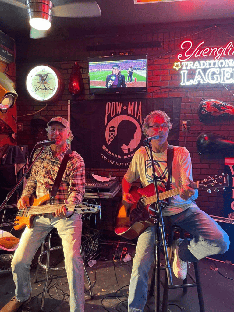 Live music performance at a cozy bar with guitarists, vibrant neon signs, and a television broadcasting a sports event.