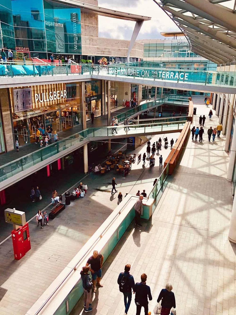 Bright indoor shopping mall with multiple store levels, shoppers, and sunlight streaming through the glass ceiling.