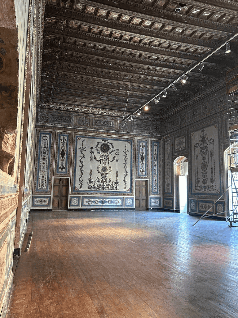 Intricate historic interior with detailed wall and ceiling decorations, elegant wooden flooring, and natural light from windows.