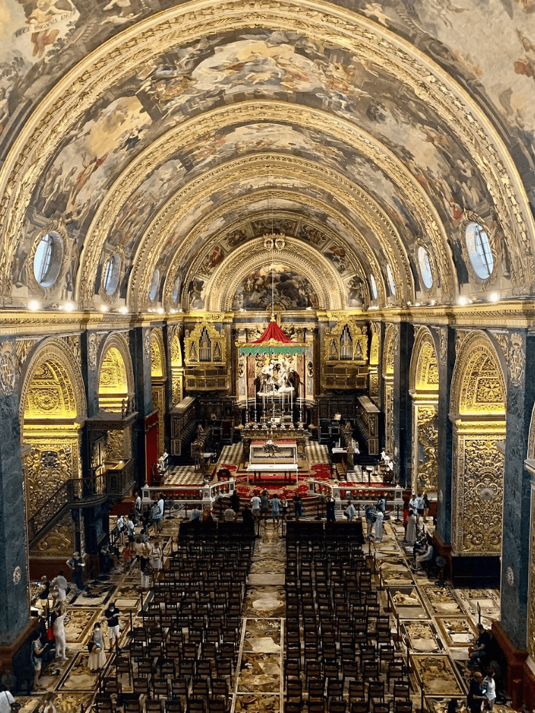Ornate church interior with baroque artwork, gold accents, and seating for visitors.
