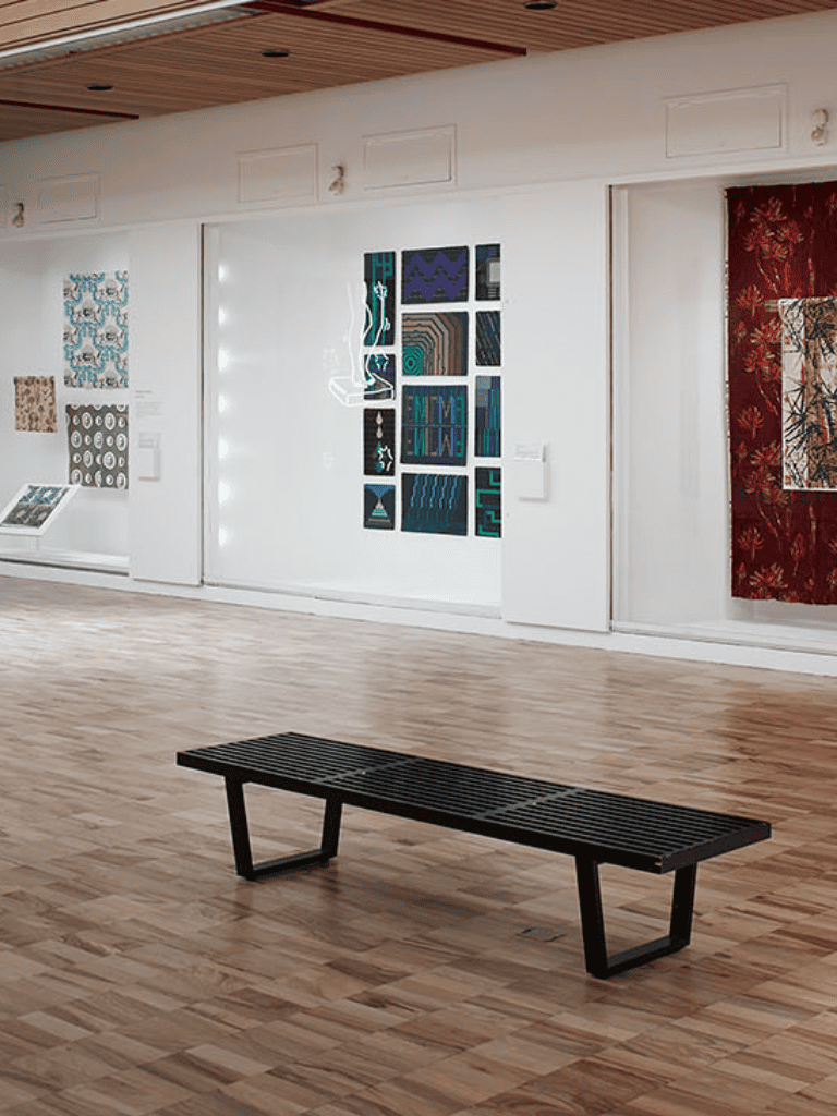 Modern art gallery with colorful textile and abstract art pieces on white walls, wooden flooring, and black bench.