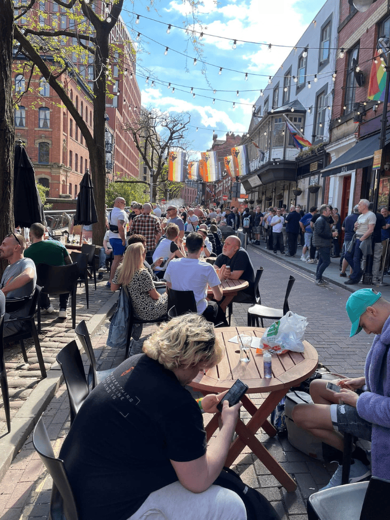 People dining outdoors on a lively city street filled with pedestrians, colorful flags, and string lights, in a vibrant urban atmosphere.