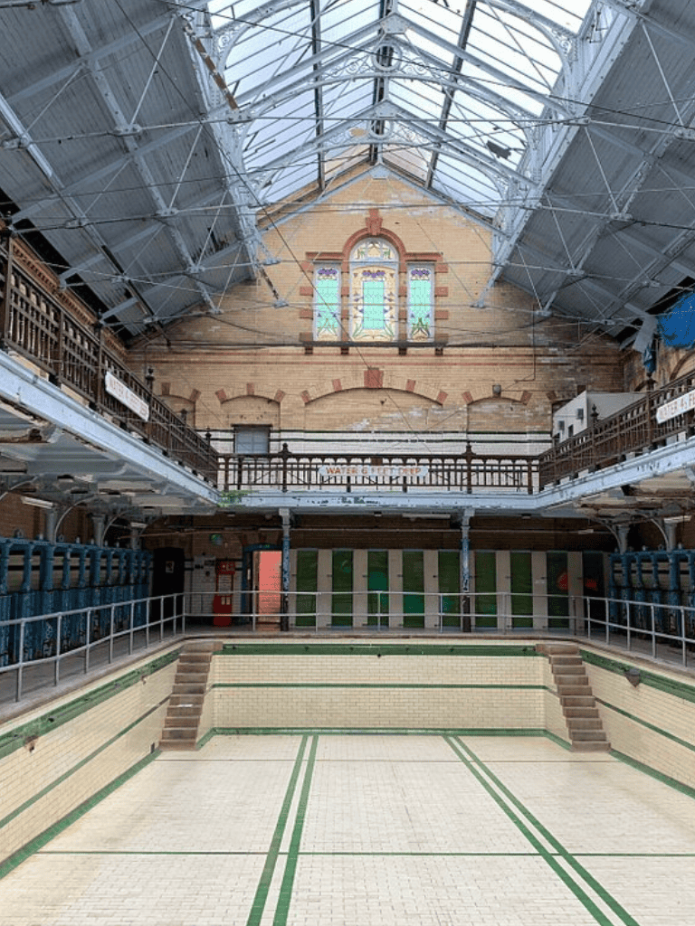 Empty vintage indoor swimming pool with high glass ceiling and brick walls, popular for historic architecture tours.