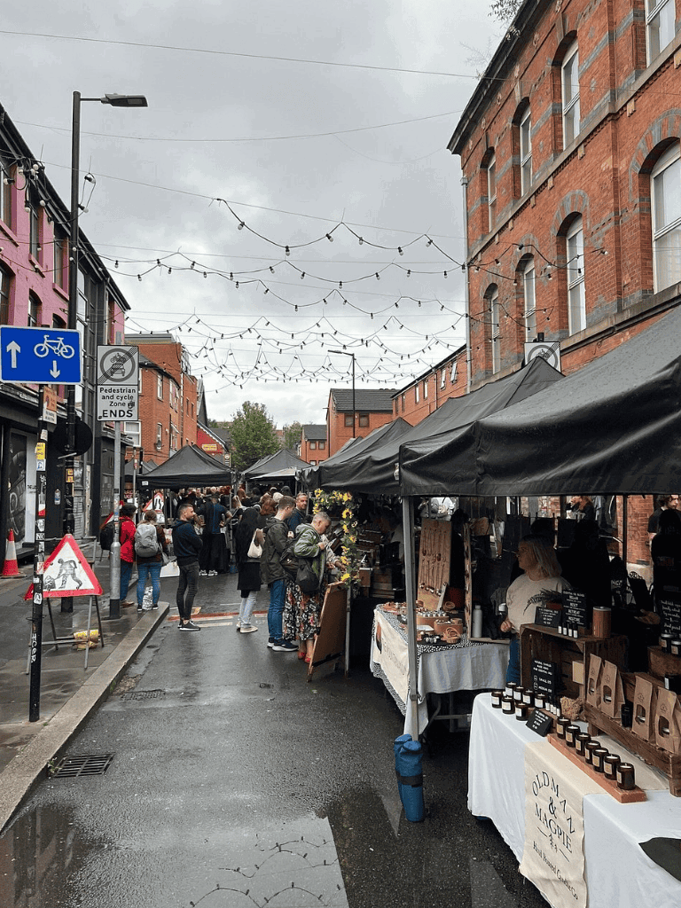 Colorful outdoor marketplace street scene with vendor stalls, shoppers, and string lights on a cloudy day, emphasizing local shopping.