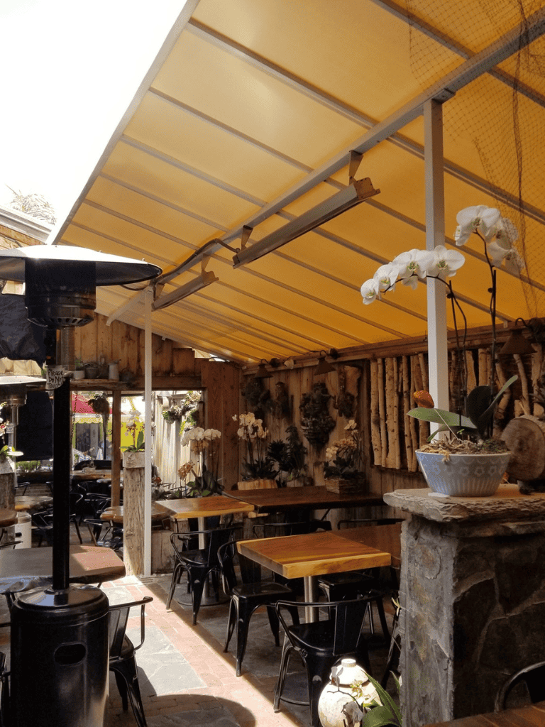 A cozy outdoor cafe with wooden tables, black chairs, and a yellow awning in a rustic setting for dining.