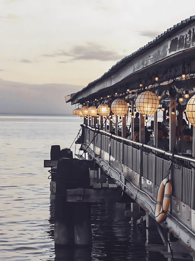 Nighttime boat restaurant on the water with warm lighting and people dining, scenic sunset view.