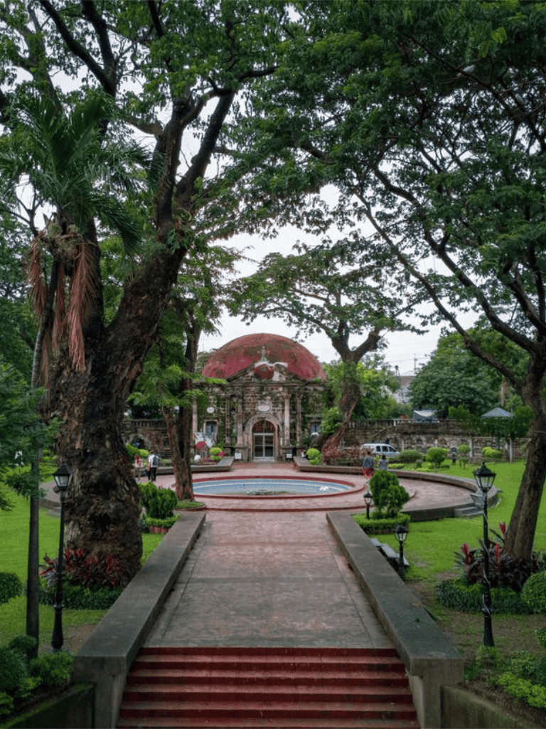 Ancient temple with lush greenery, stone pathway, and fountain, surrounded by tall trees and garden lamps.