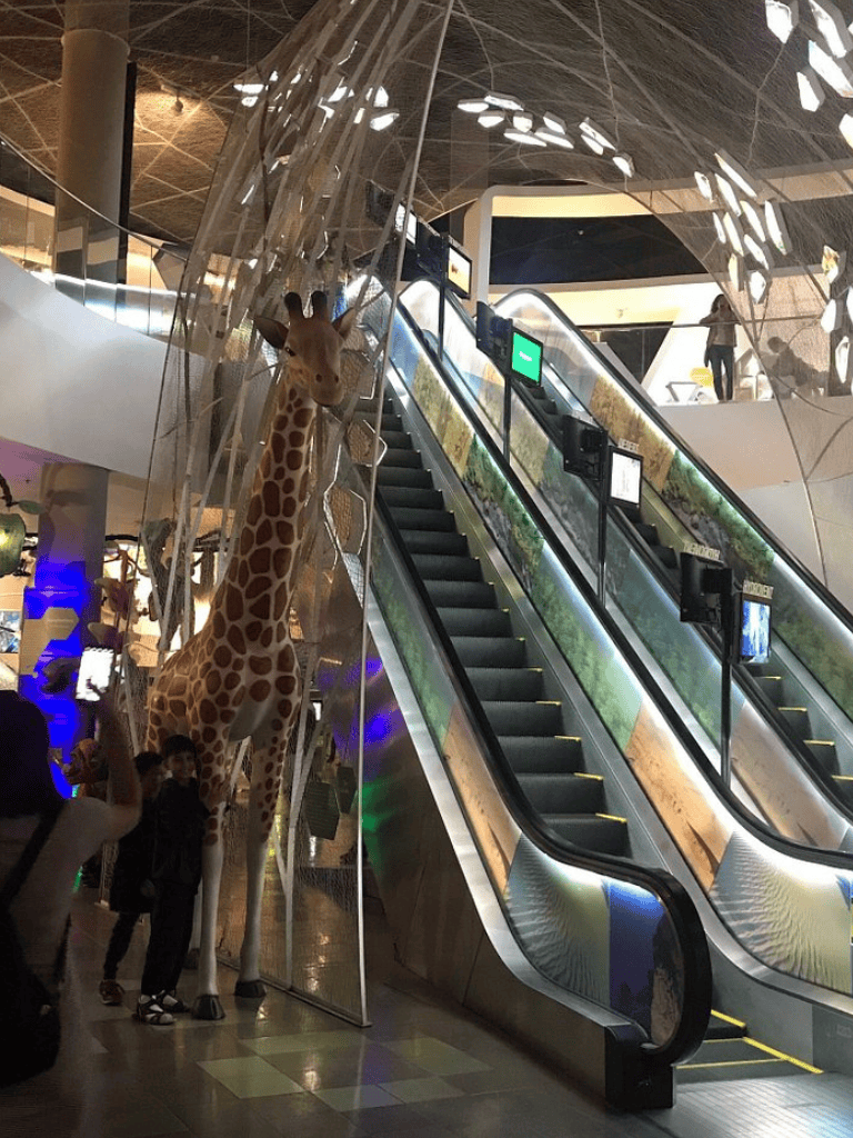Giraffe statue at shopping mall with escalators and modern architecture in the background.