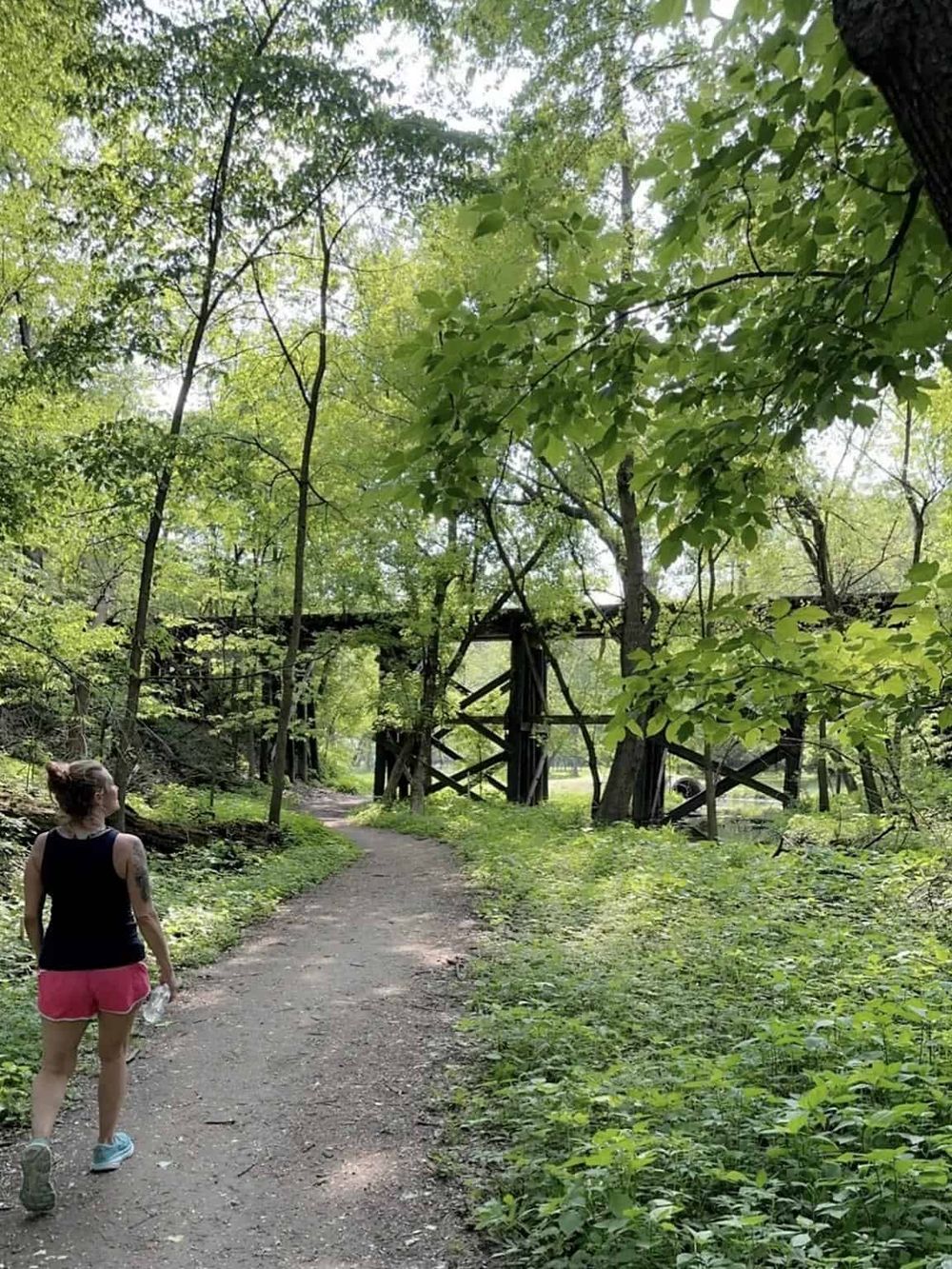 Lush green forest trail with a woman in casual attire walking towards an old wooden bridge. Perfect for nature walks and outdoor adventures.
