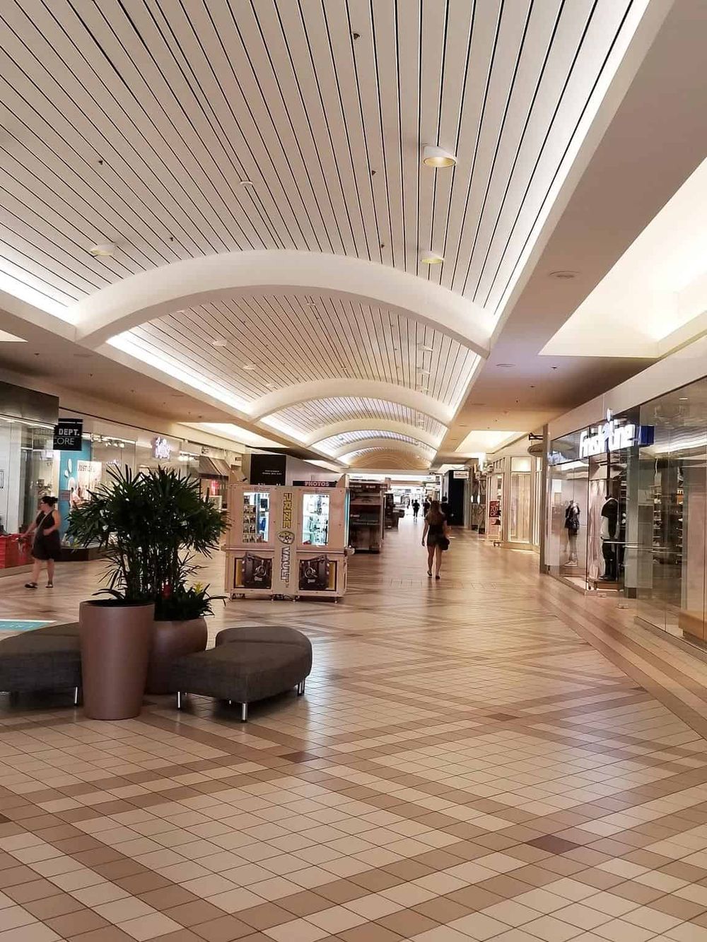 Bright indoor shopping mall corridor with shops, potted plant, and seating area for travelers.