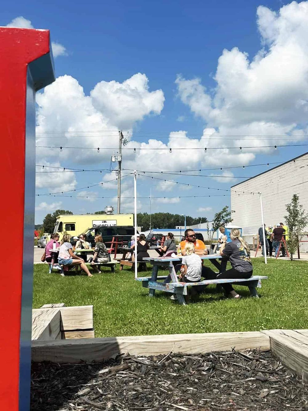 Outdoor dining at a casual food park, family and friends enjoy meals on sunny day under string lights.