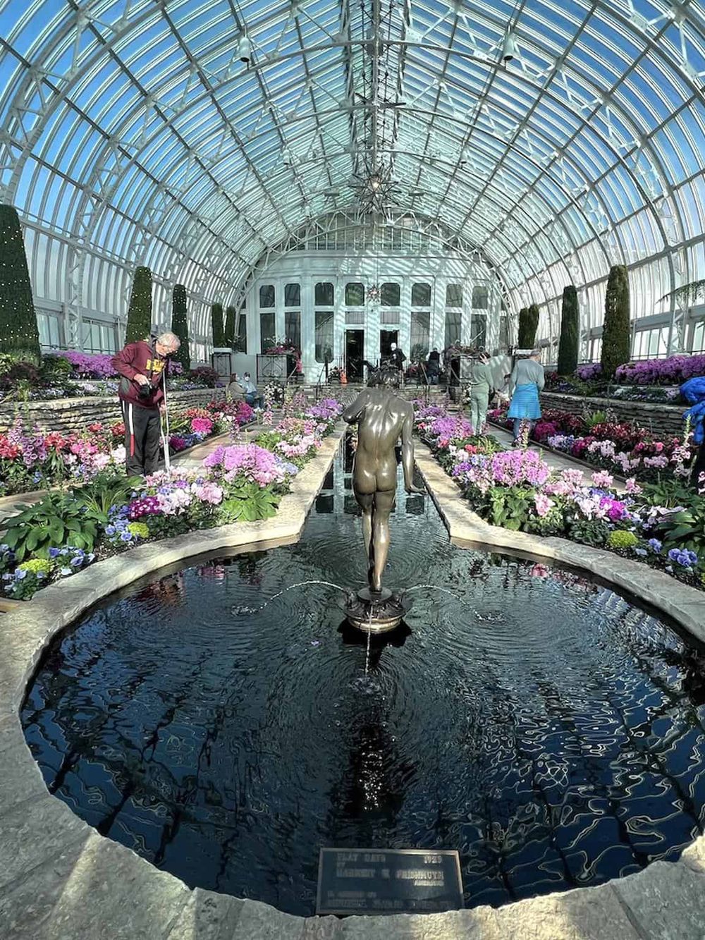 Colorful flower exhibit inside a glass conservatory with a fountain and statues, part of Quest for Directions botanical garden.