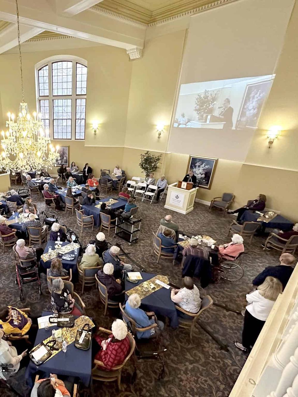 Aerial view of a conference room with attendees listening to a speaker at a seminar, elegant chandelier, and large windows.