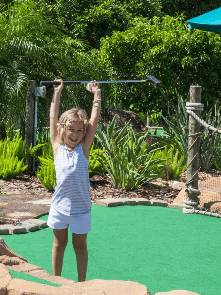 Child playing mini golf outdoors, smiling and holding golf club.