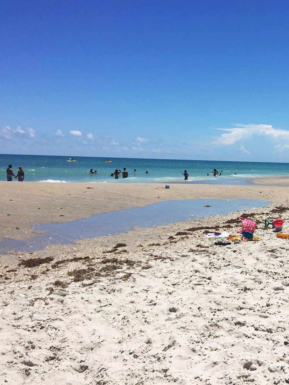 Bright sandy beach with families enjoying the ocean, clear blue sky, and water sports activities, perfect for navigation and travel planning.