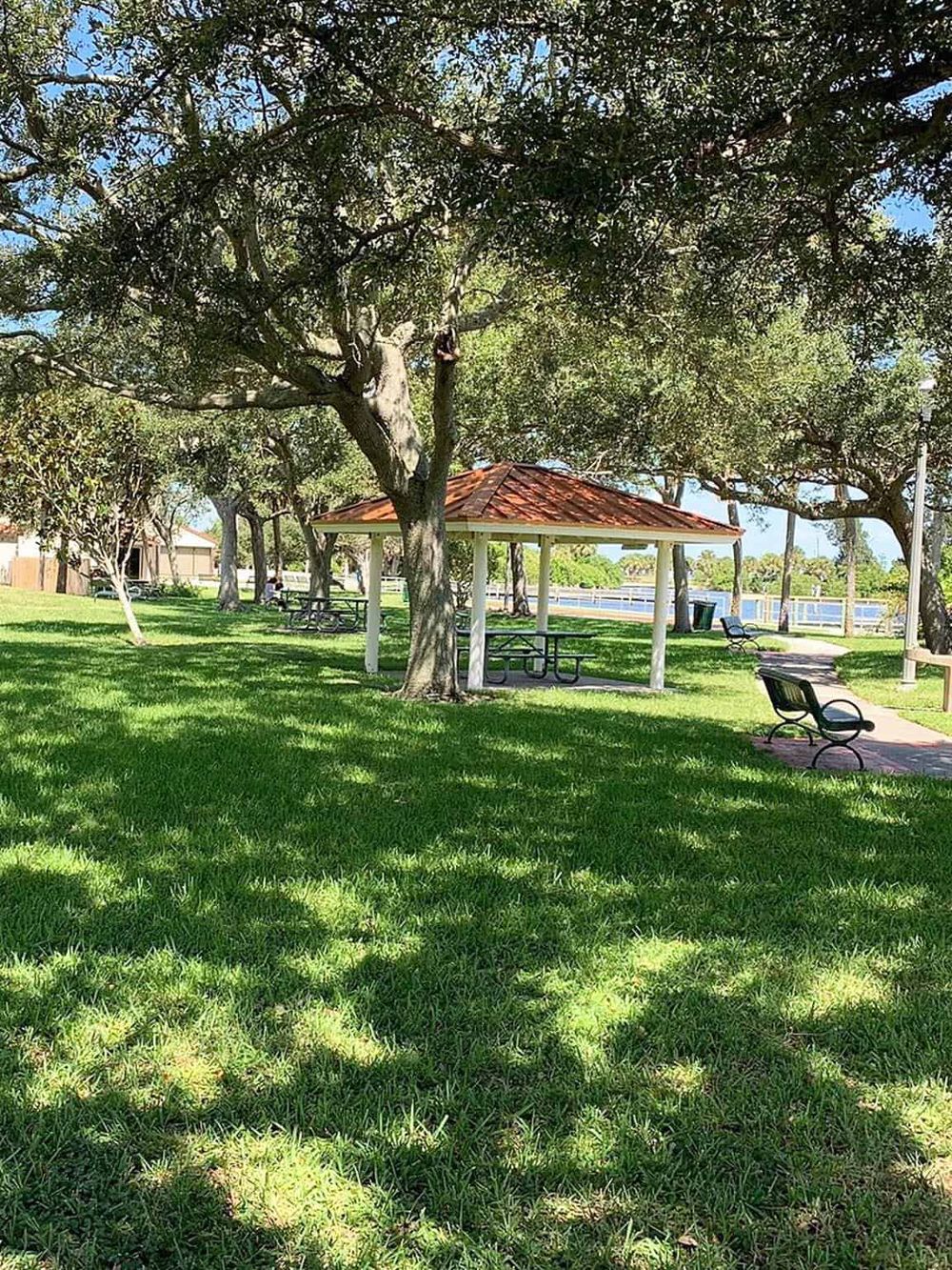 Shady park with trees, picnic shelter, benches, and a waterfront view, ideal for relaxation and outdoor activities.