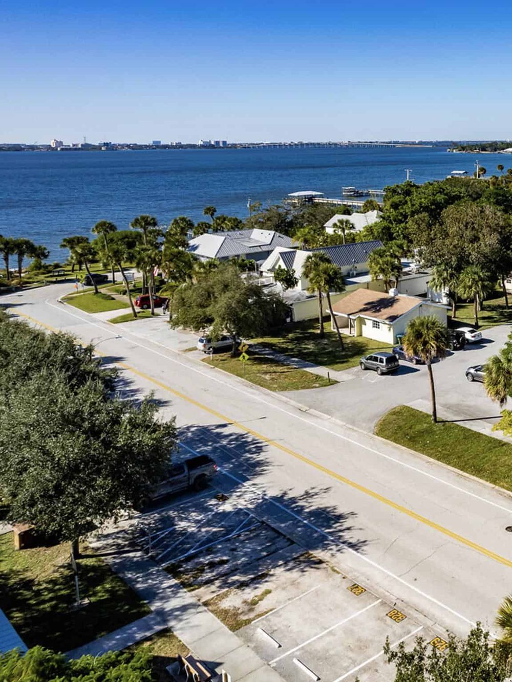 Waterfront neighborhood with palm trees, houses, and parking, overlooking a large river or bay.