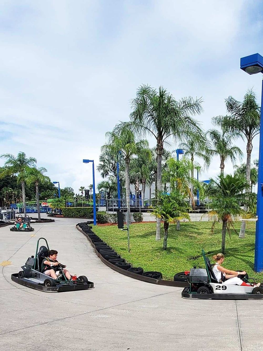 Racetrack with children driving go-karts surrounded by palm trees and blue light poles.