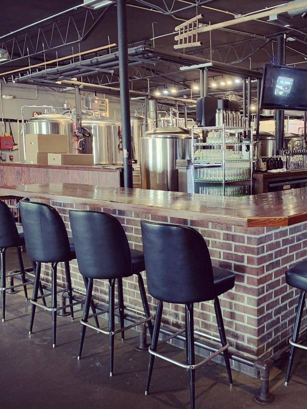 Industrial brewery bar setup with brewing tanks, glassware, and seating for tasting beers.